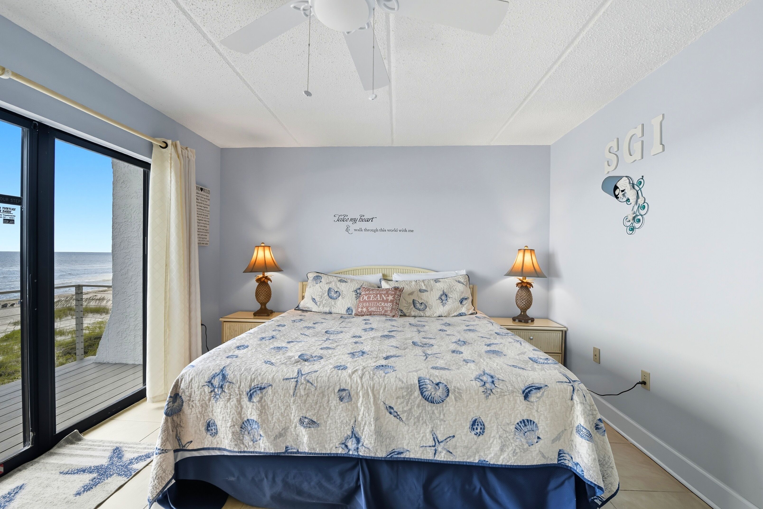 Ein Schlafzimmer mit einem Bett, zwei Nachttischlampen, einem Deckenventilator, Meerblick und einer Wand mit einem dekorativen Schild.