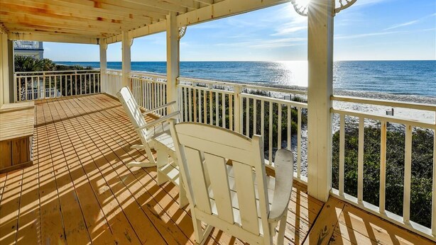 Une terrasse en bois avec des chaises blanches et une table, donnant sur une plage et agrémentée d’une rambarde.