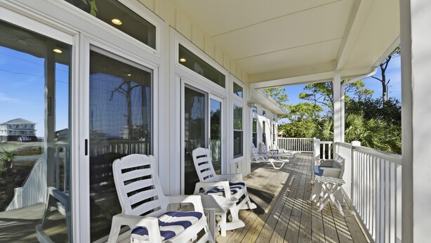 A covered deck with white chairs and a table, large glass doors, and a view of the ocean and a house. - WELCOME to our house, HEATWAVE for an unforgettable beach experience on beautiful St. George Island. (St. George Island)