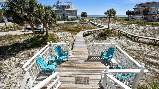 A wooden boardwalk leading to a beach house with a balcony and blue chairs.