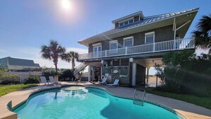 A swimming pool with a sunlit deck area, a covered outdoor seating area, and a two-story building with a balcony.