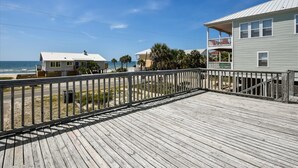 A wooden deck with a railing overlooking a beach, a building with a balcony, and a clear view of the ocean.