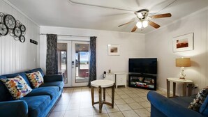 A living room with a blue sofa, a round wooden table, a ceiling fan, a TV, and a small side table with a lamp.