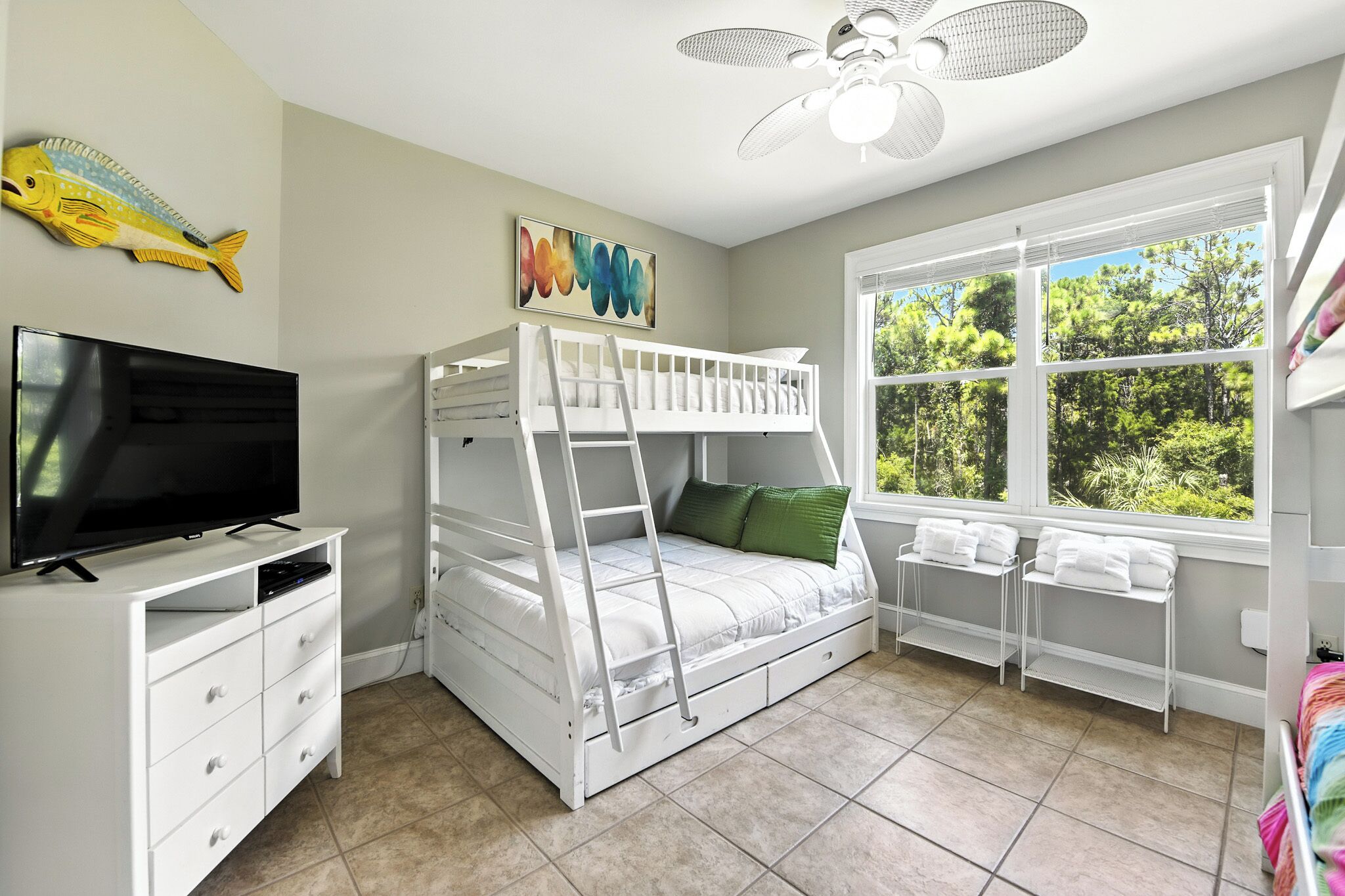 A bedroom with a bunk bed, a TV, a ceiling fan, a painting, and a window with a view of trees.
