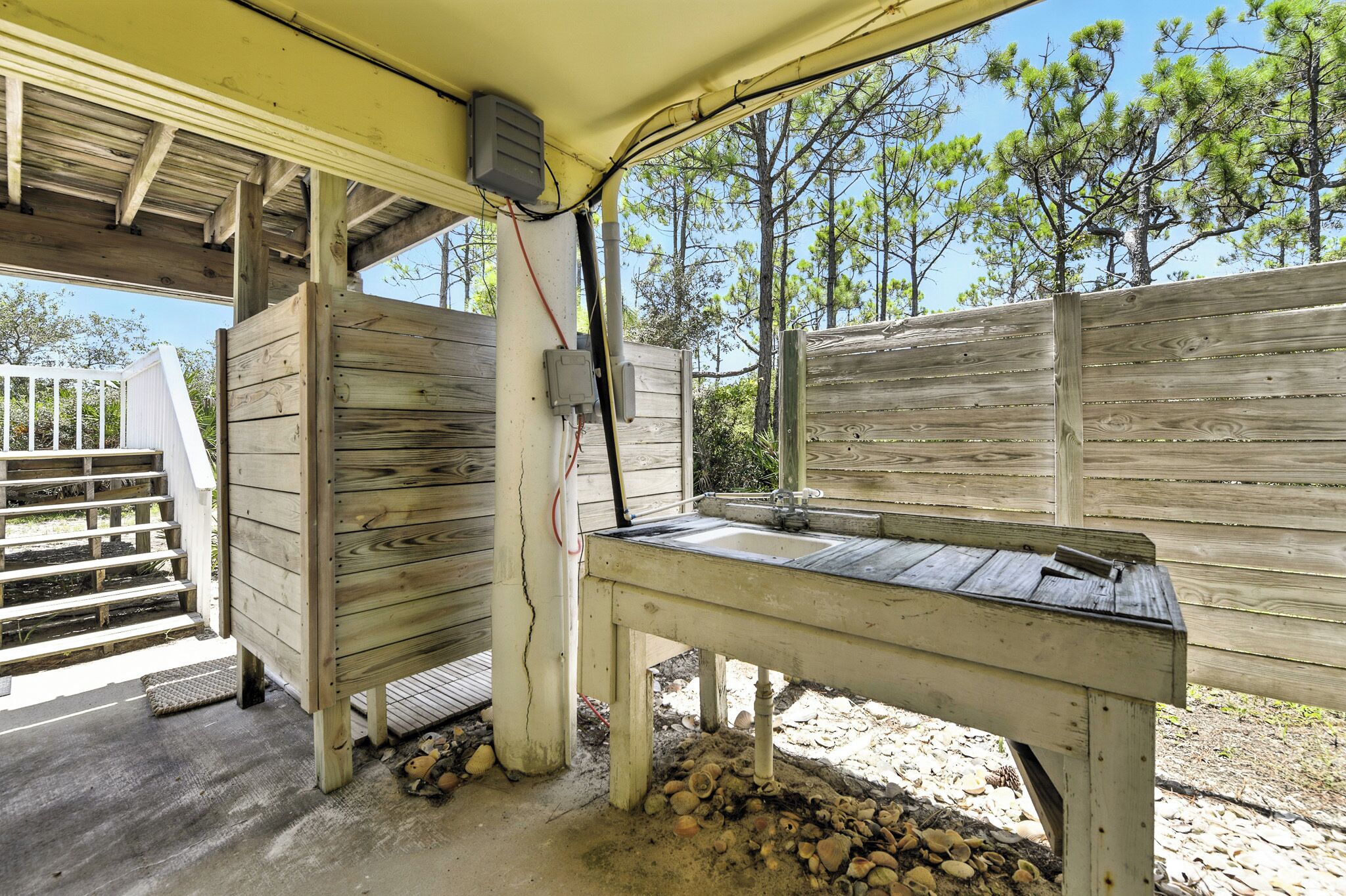 Un banc en bois placé à l’extérieur, sous une zone abritée, avec une clôture en bois et des marches menant à un bâtiment blanc.