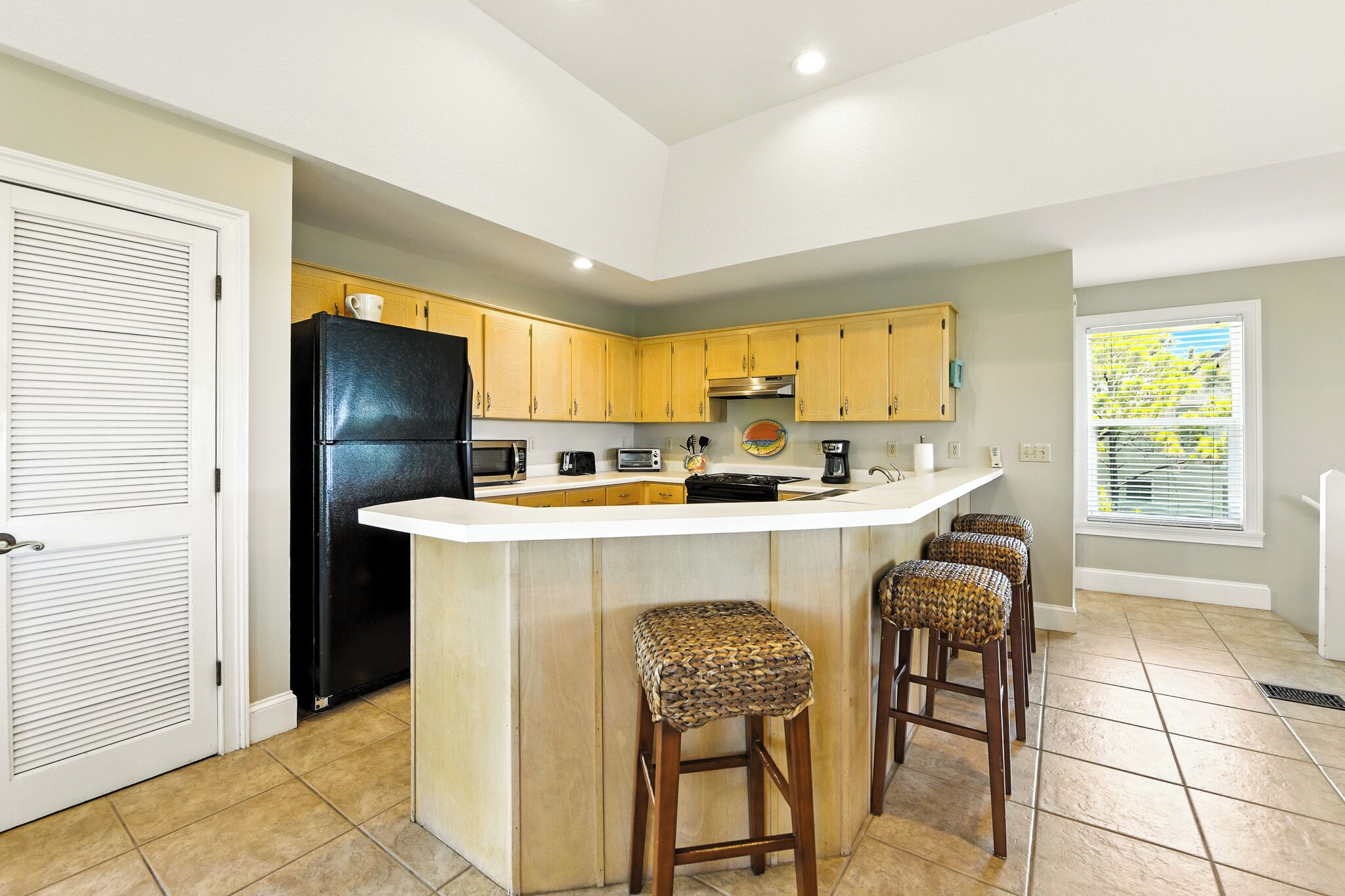 A kitchen with a central island, wooden cabinets, a black refrigerator, and a window with blinds.
