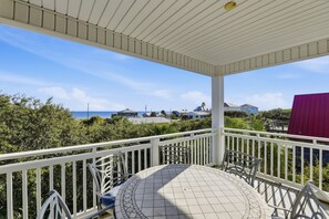 A covered outdoor area with a tiled floor and white railings, overlooking a coastal landscape with houses and greenery. - THE MINUTE YOU STEP INTO A WAVE FROM IT ALL, you understand the name! (St. George Island)