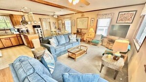 A living room with a sofa, coffee table, and chairs, a kitchen area with cabinets and appliances, and a dining table with chairs.