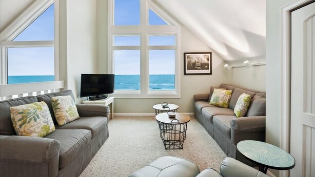 A cozy living room with a large window overlooking the ocean, a flat-screen TV, two round tables, and a sofa with floral cushions.