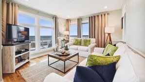 A modern living room with a television, sofa, coffee table, and large windows offering a view of the sea.