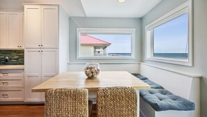 A kitchen with white cabinets, a wooden table, wicker chairs, and a view of the ocean through a window.