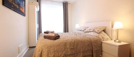 A bedroom with a bed, a nightstand with a lamp, and a window with curtains.