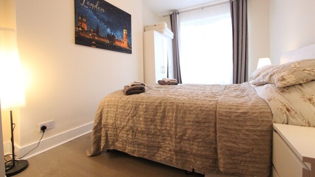 A neatly made bed with a beige quilt, a night sky poster on the wall, and a window with curtains. - Cosy One Bedroom Apartment- Marble Arch (London)