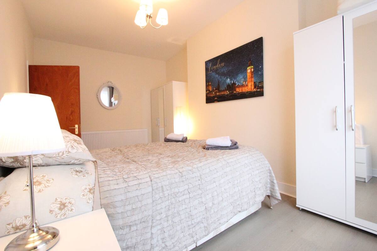A bedroom with a bed, a bedside table with a lamp, a wardrobe, a round mirror, and a wall with a night-time cityscape painting.