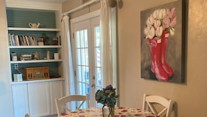 A dining room with a floral tablecloth, white chairs, a vase with flowers, and a bookshelf with books and decorative items.