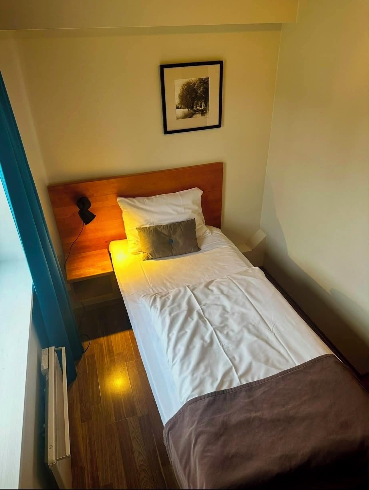 A small, single-bed room with a wooden headboard, a framed picture on the wall, and a window with curtains.