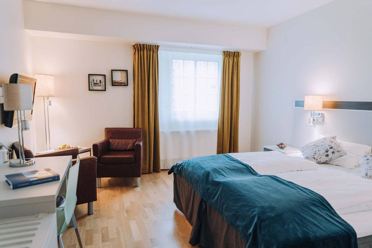 A hotel room with a bed, a desk with a lamp, a chair, and a window with curtains.