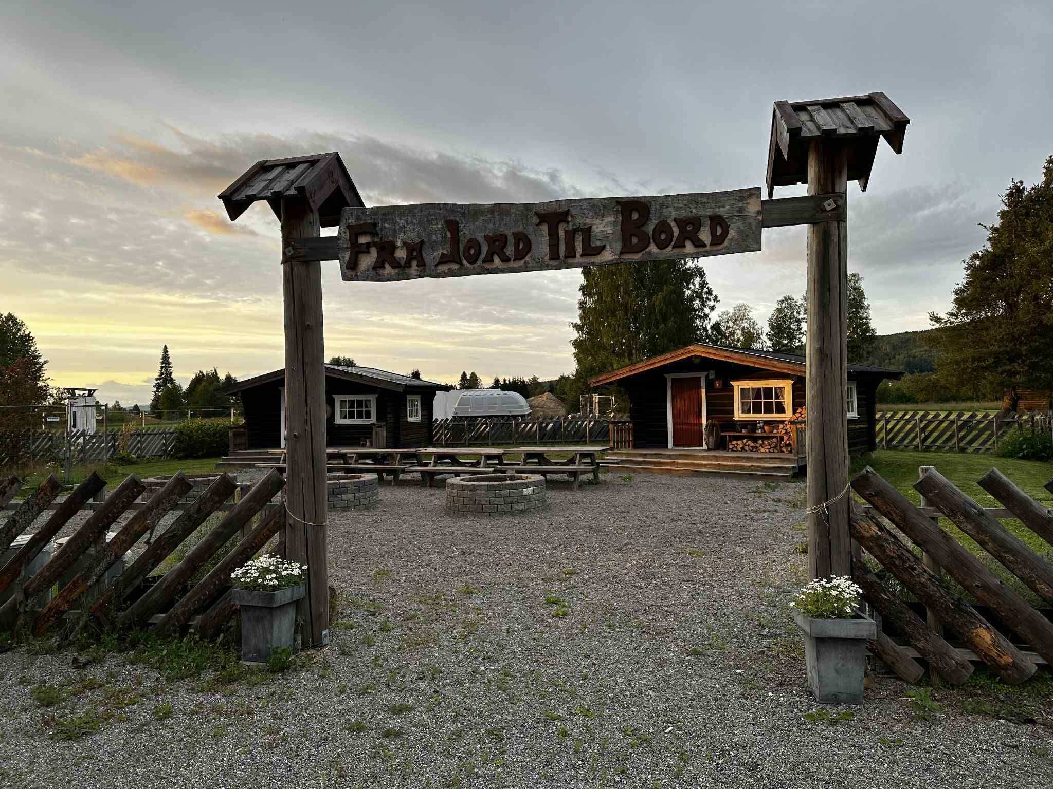 A wooden sign with the text 'FRA LORD TIL BORD' and a gravel path leading to a building with a covered porch.