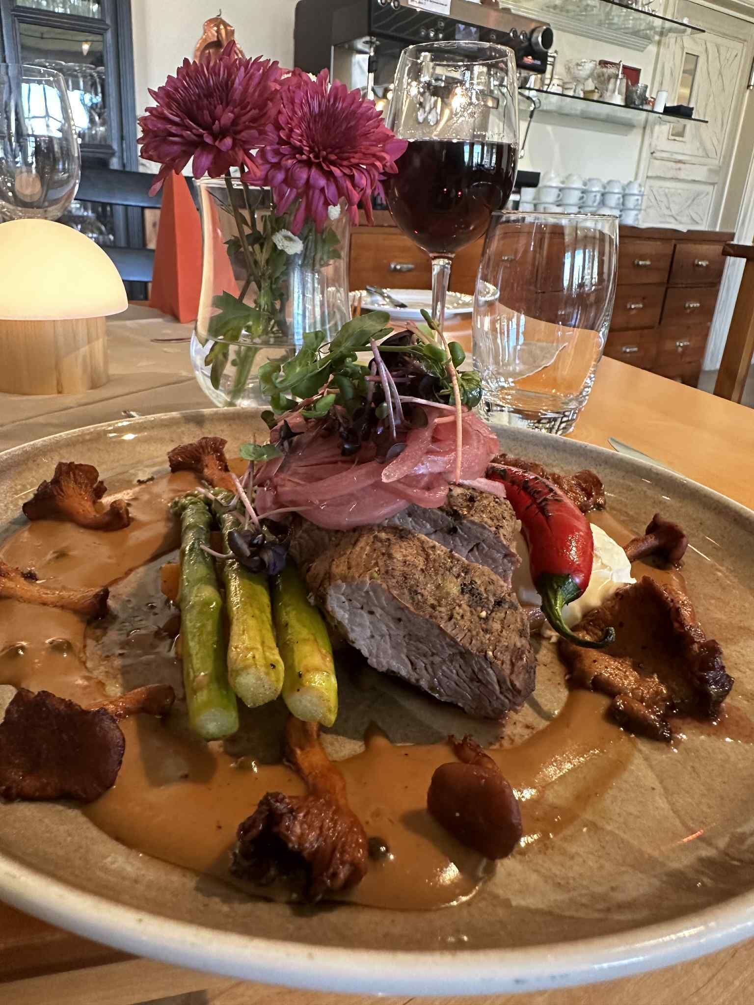A plated meal with various meats and vegetables, a glass of red wine, and a vase with flowers.