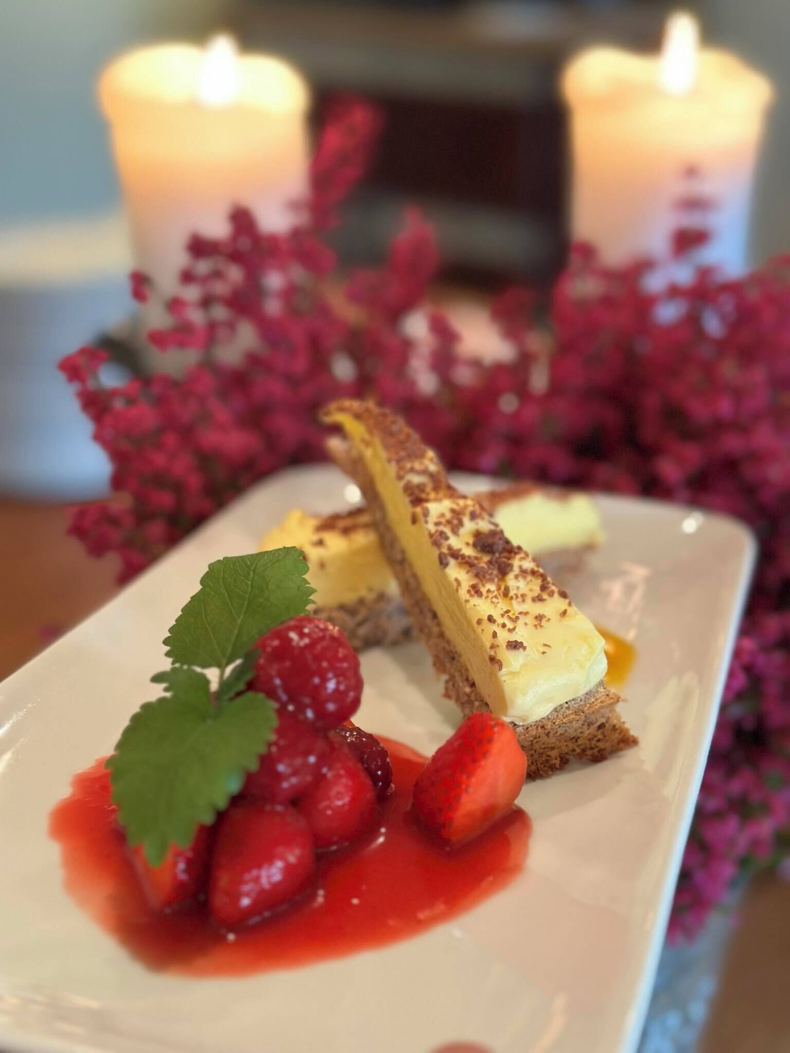 Een taartbord met een stuk cheesecake op een cracker, versierd met een aardbei en een blaadje, met rode saus en bessen, voor een achtergrond met roze bloemen en kaarsen.