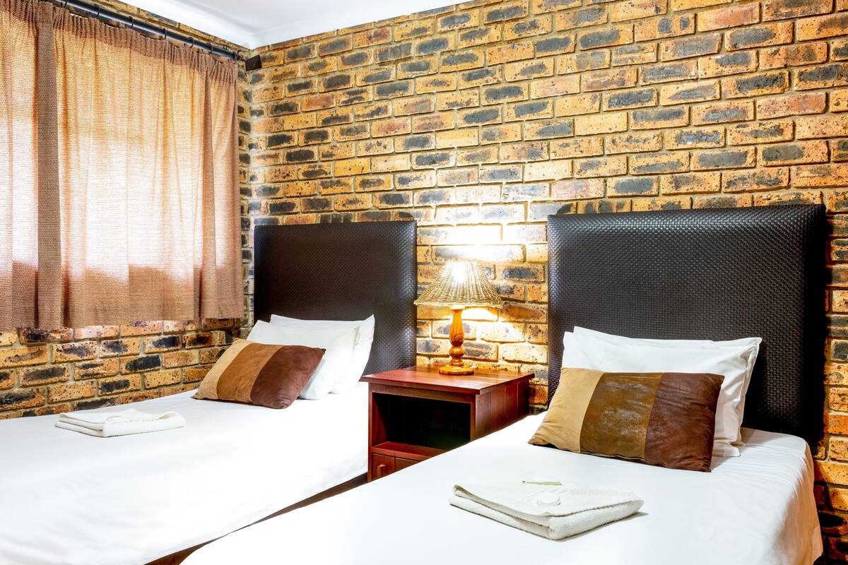A hotel room with two beds, a brick wall, a window with curtains, and a nightstand with a lamp.