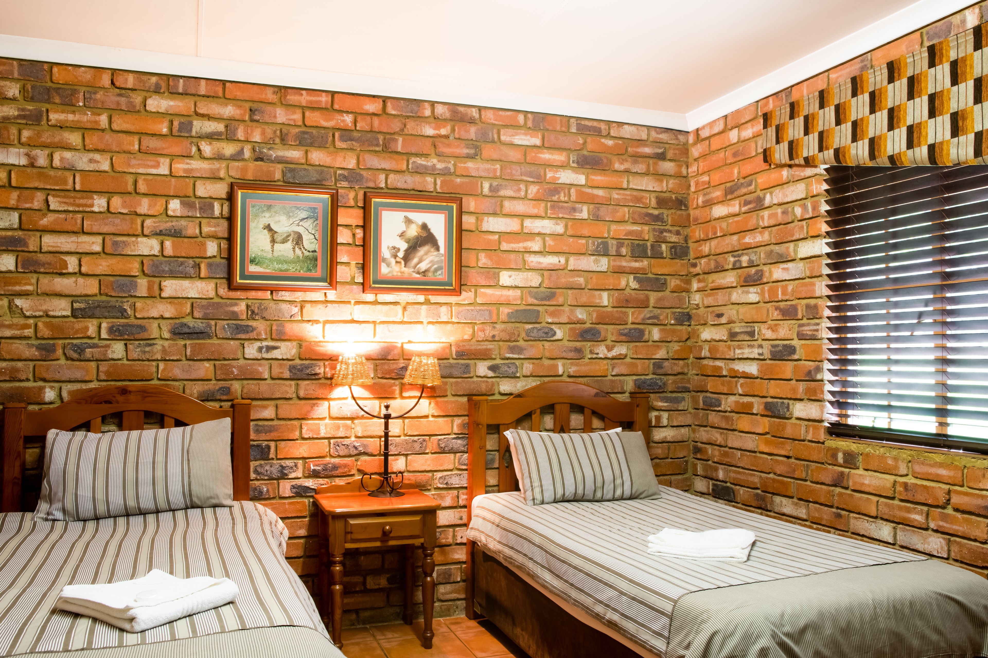 A room with two beds, a wooden headboard, a nightstand, a lamp, and a brick wall with framed pictures.