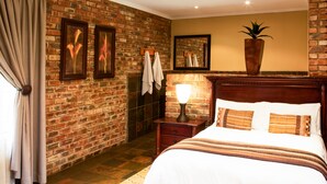 A hotel room with a brick wall, a bed with white linens, a nightstand with a lamp, and a large potted plant. - Elgro River Lodge (Potchefstroom)