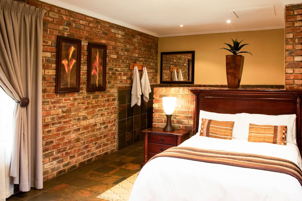 A hotel room with a brick wall, a bed with white linens, a nightstand with a lamp, and a large potted plant.