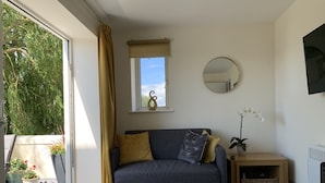 Un salon avec un canapé gris, un tapis à motifs, une petite table sur laquelle est posée une plante, une fenêtre avec des rideaux et un miroir fixé au mur.