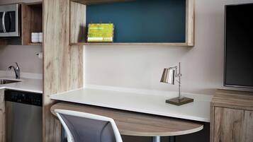 Una cocina moderna con microondas integrado, un lavaplatos y un lavamanos. Hay una mesa redonda con una silla, un estante pequeño con una libreta y un televisor montado en la pared.
