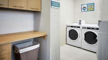 A laundry room with two washing machines, a drying machine, a wooden shelf, a trash bin, and signs indicating laundry products are available and to wipe the front desk.