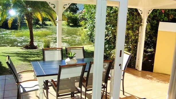 A glass-enclosed patio with a table and chairs, a potted plant, and a view of a garden with a palm tree. - River Club 4213 (Plettenberg Bay)