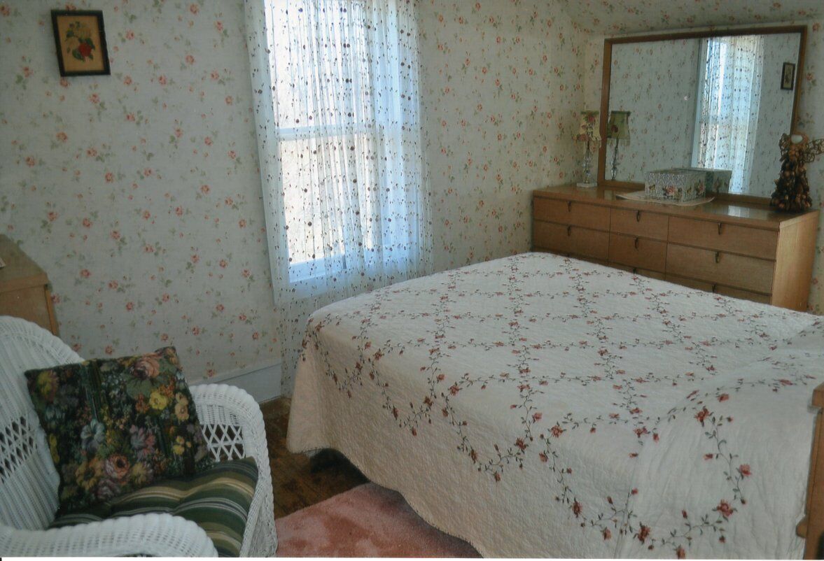 A bedroom with a floral-patterned bedspread, a wooden dresser, a wicker chair, and a window with sheer curtains.
