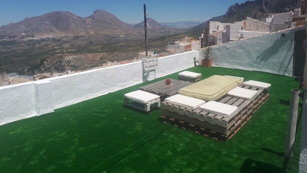 Une terrasse sur le toit offrant une vue sur les montagnes, un mur blanc et un espace recouvert d’une pelouse artificielle verte, avec une table et des chaises.