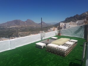 Une terrasse sur le toit offrant une vue sur les montagnes, un mur blanc et un espace recouvert d’une pelouse artificielle verte, avec une table et des chaises.