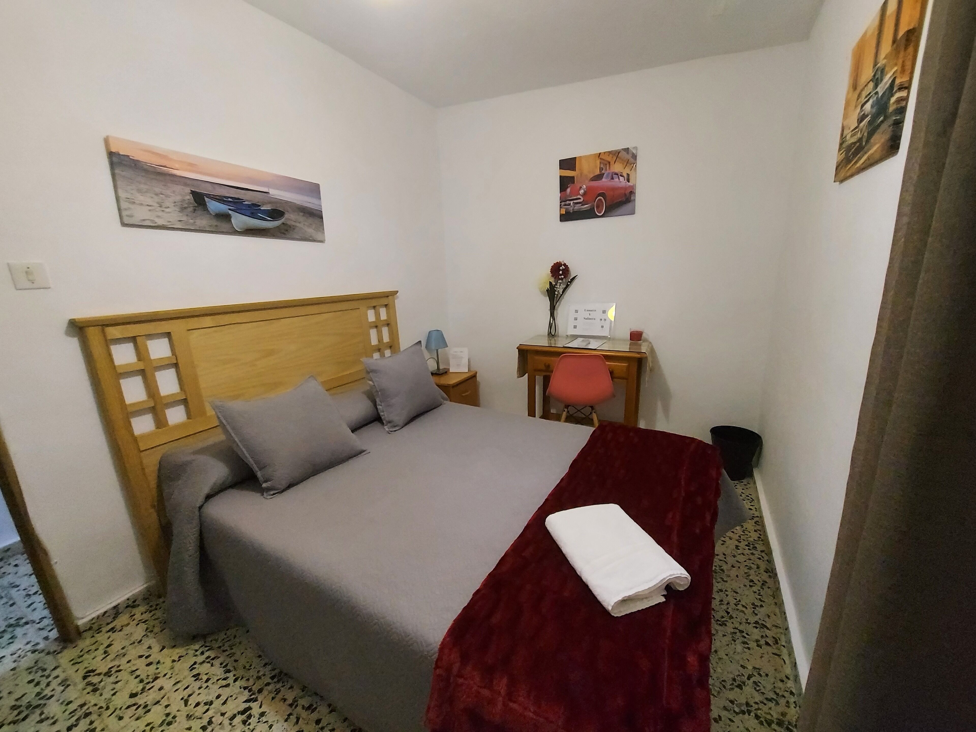 Una cama bien hecha con cabecera de madera, un escritorio con una silla y dos cuadros en las paredes.