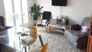 Un salon avec une table en verre, des chaises jaunes, un canapé gris, un téléviseur et une bibliothèque contenant des magazines.