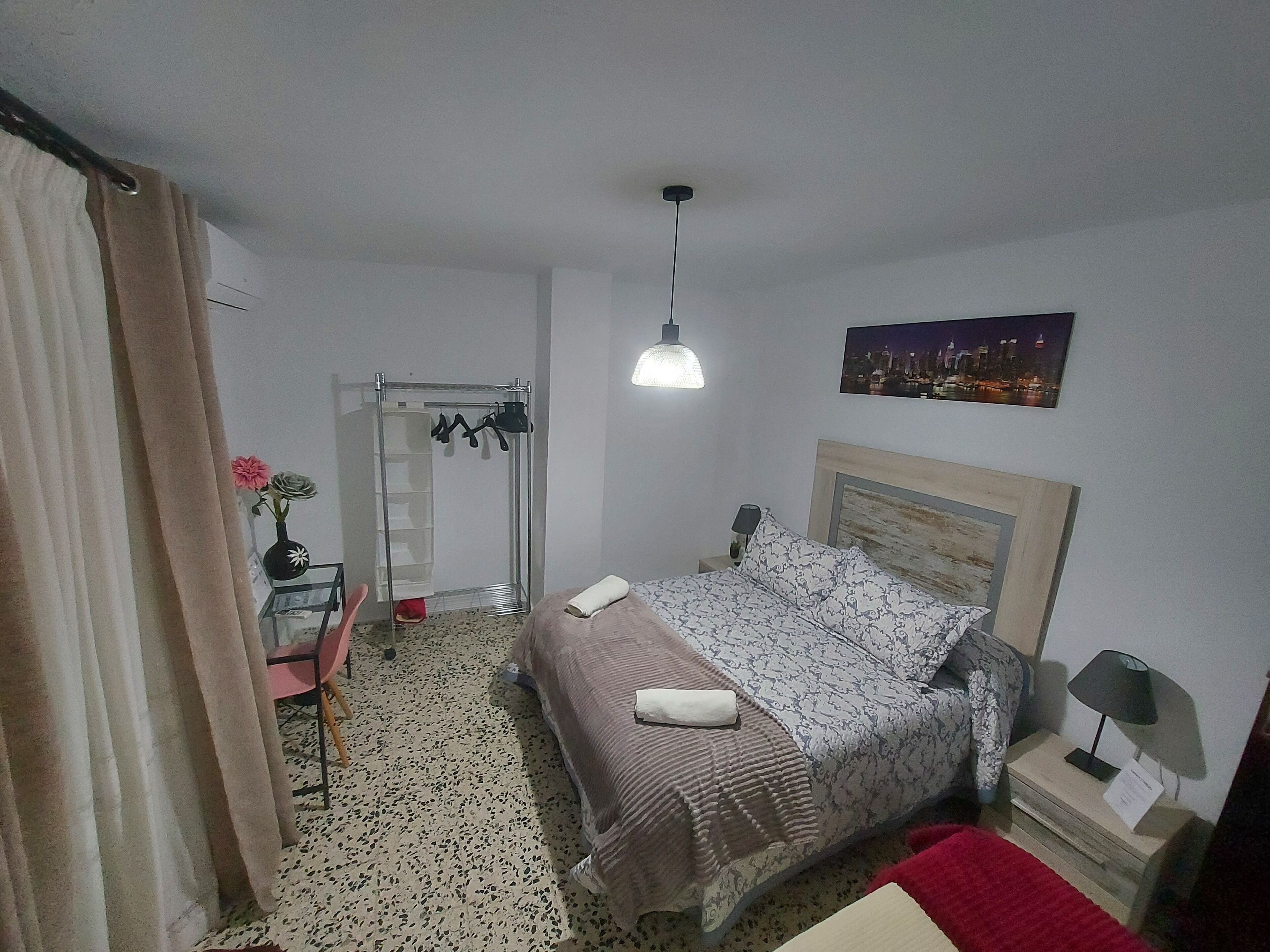 Un dormitorio con cama, mesita de noche, lámpara, un cuadro y un tendedero.