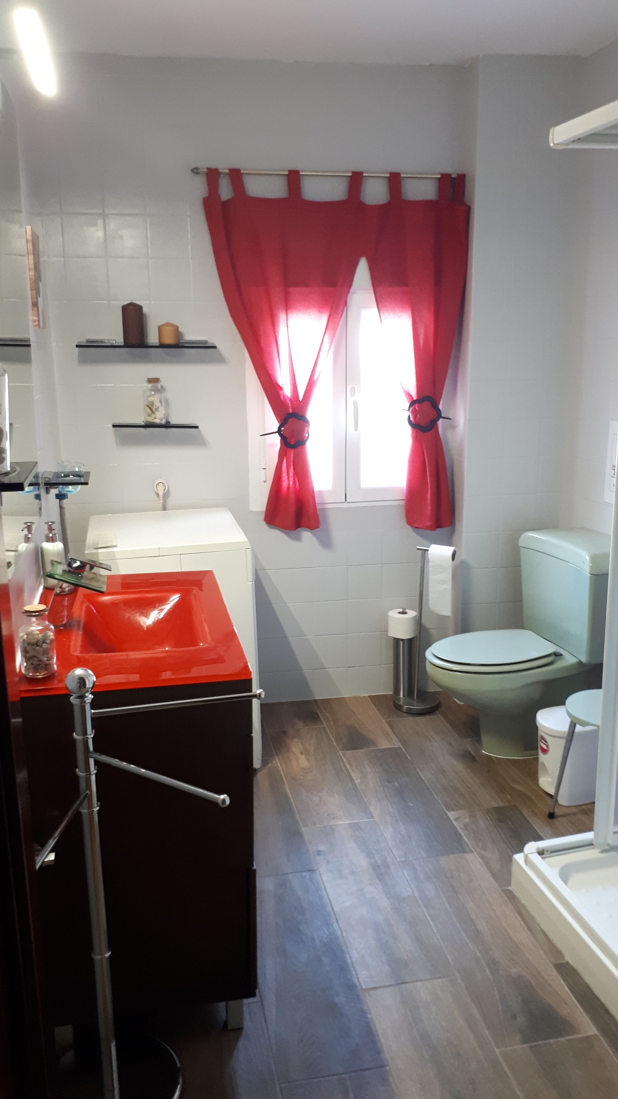 Une salle de bain avec un lavabo, des toilettes et une douche, le tout de couleur rouge. On y trouve des étagères avec des articles de toilette et un porte-serviettes.