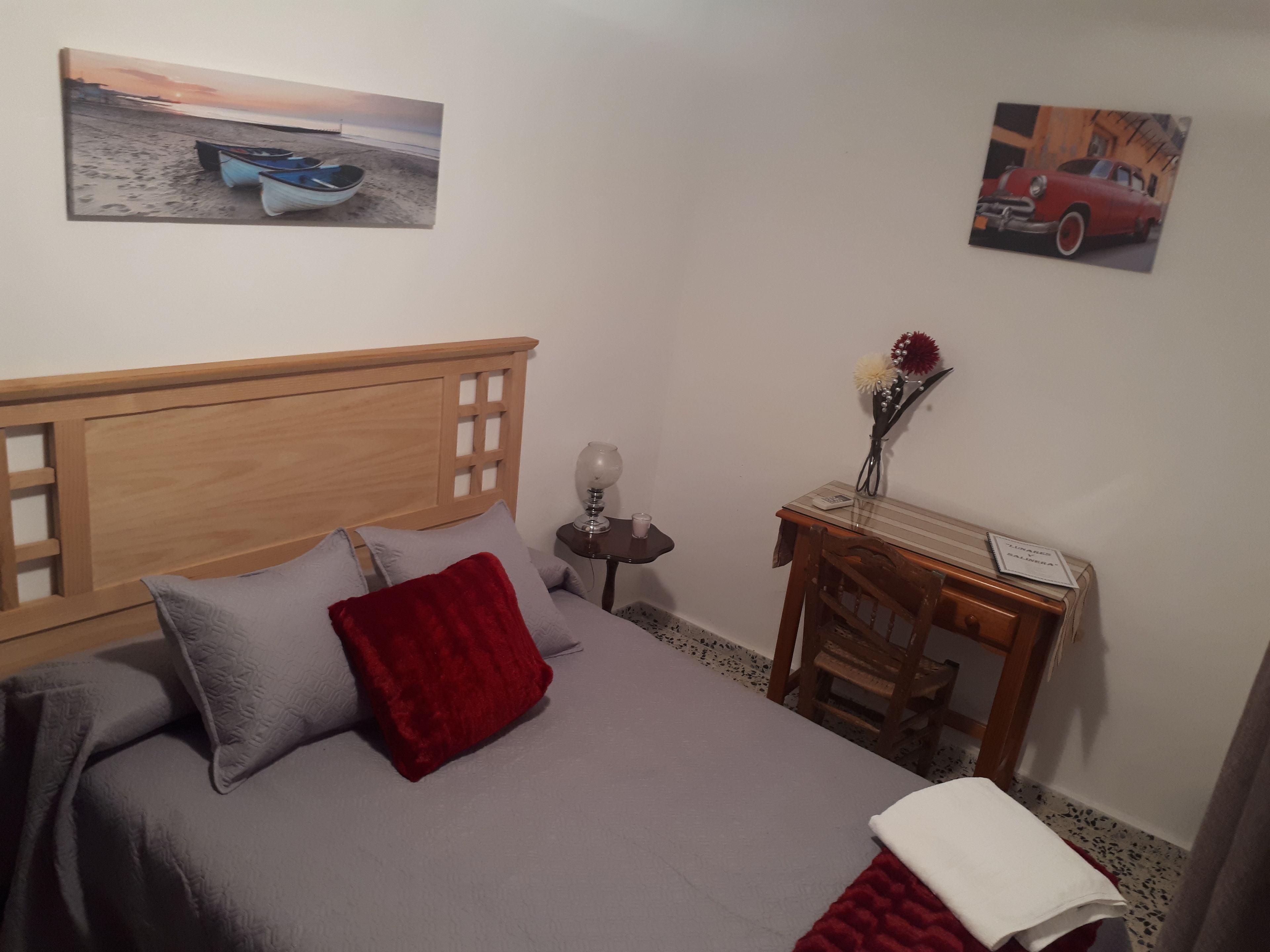 Une chambre à coucher avec une tête de lit en bois, un lit avec une literie grise et rouge, une table de chevet avec une lampe, une petite table avec un vase de fleurs et deux œuvres d’art murales.