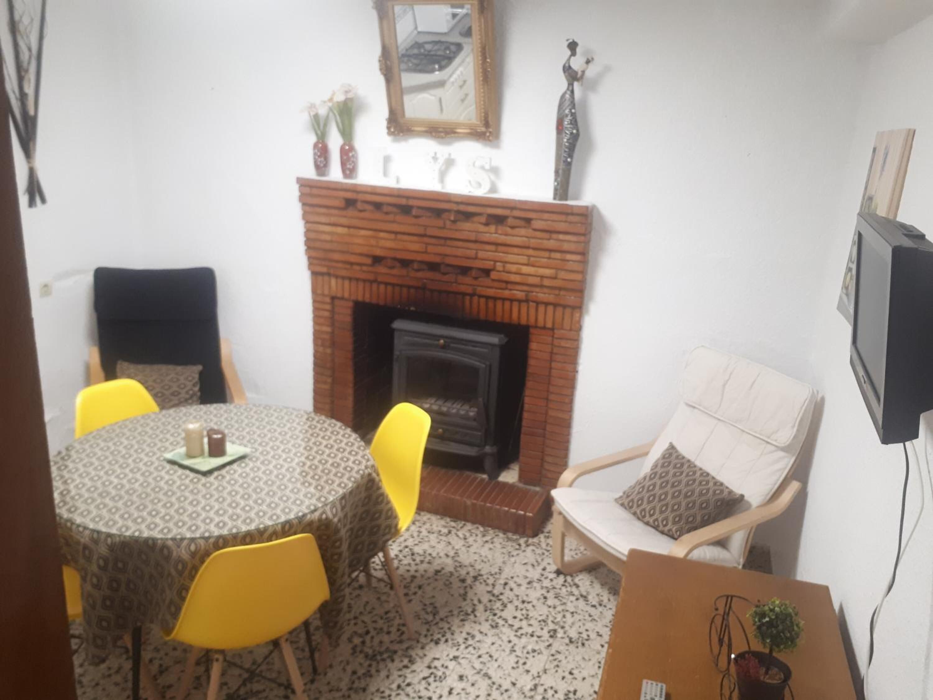 Een gezellige woonkamer met een houten haard, een ronde tafel met gele stoelen, een bank met een gekleurd kussen, en een televisie aan de muur.
