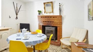 Une pièce avec une cheminée en briques, une table ronde entourée de chaises jaunes, un fauteuil blanc et une petite bibliothèque.