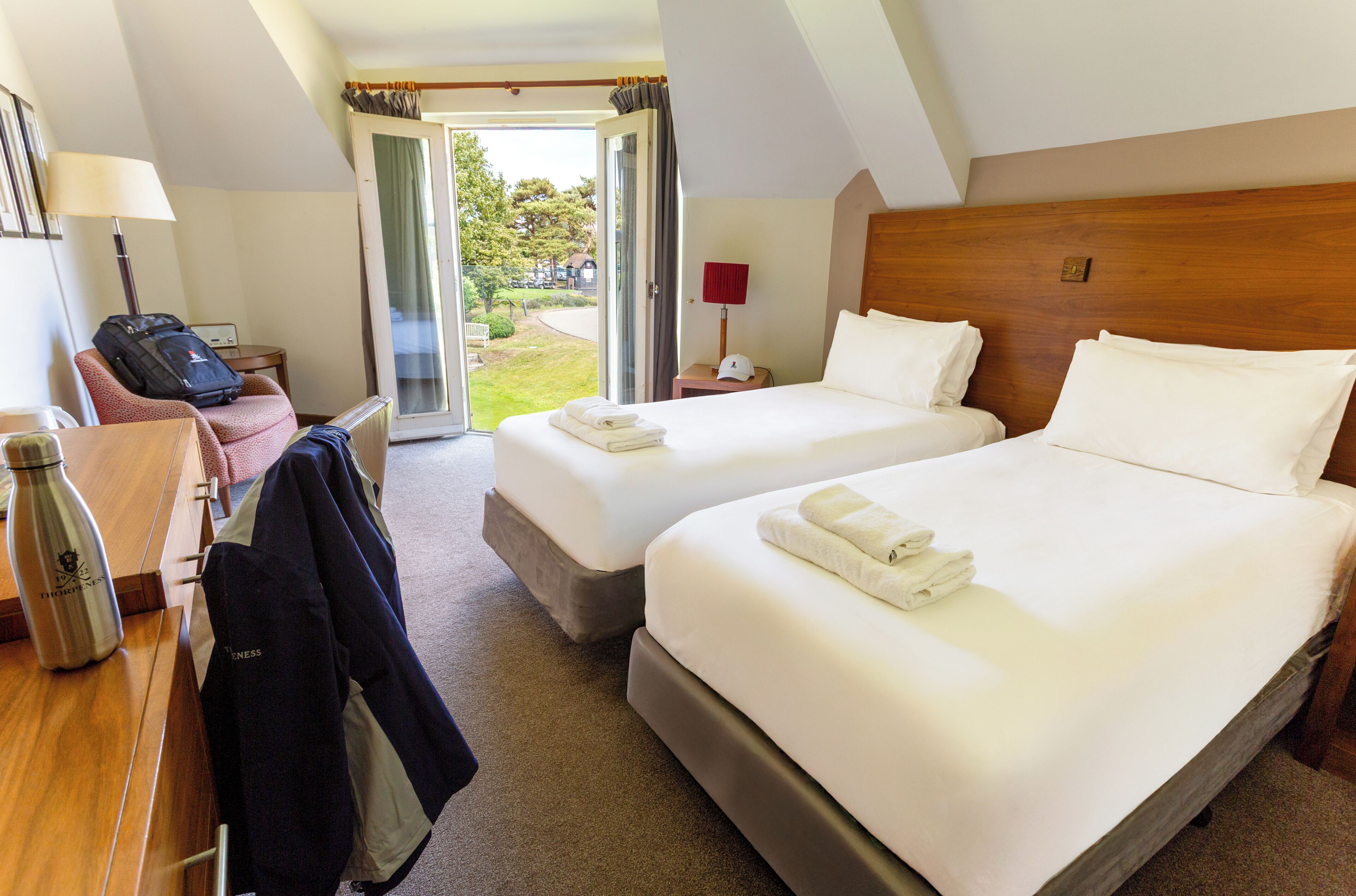 A hotel room with two beds, a desk with a lamp, a chair, a suitcase, and a view of the outdoors.