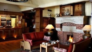 A cozy living room with a brick fireplace, red sofas, and a bar area in the background. - Thorpeness Golf Club and Hotel (Leiston)