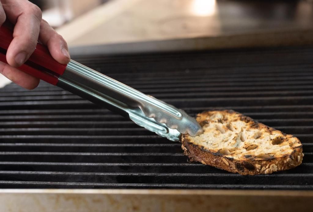 A hand using tongs to place a slice of bread on a grill.