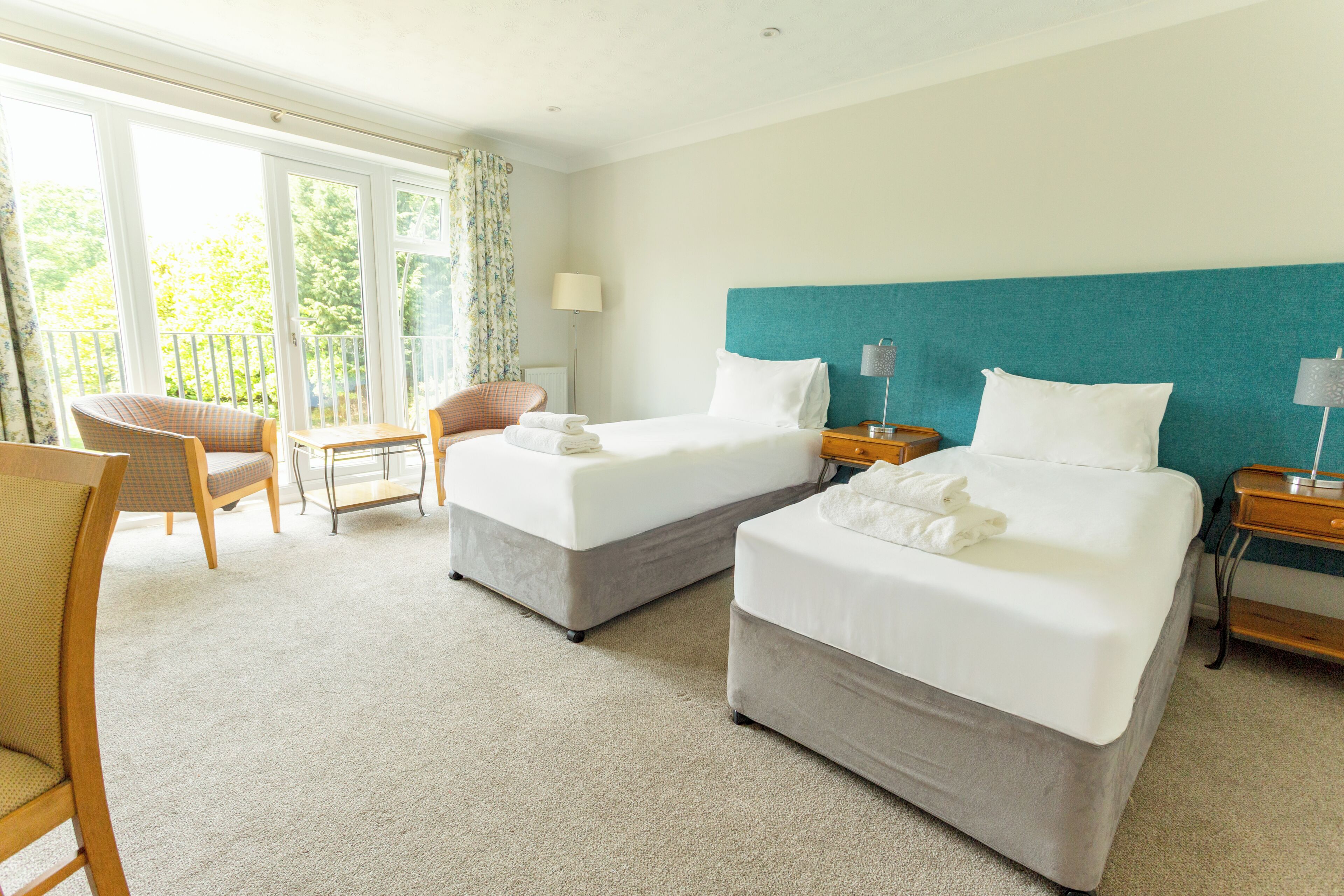 A hotel room with two beds, a sitting area with a chair, a lamp, and a view of the outdoors.