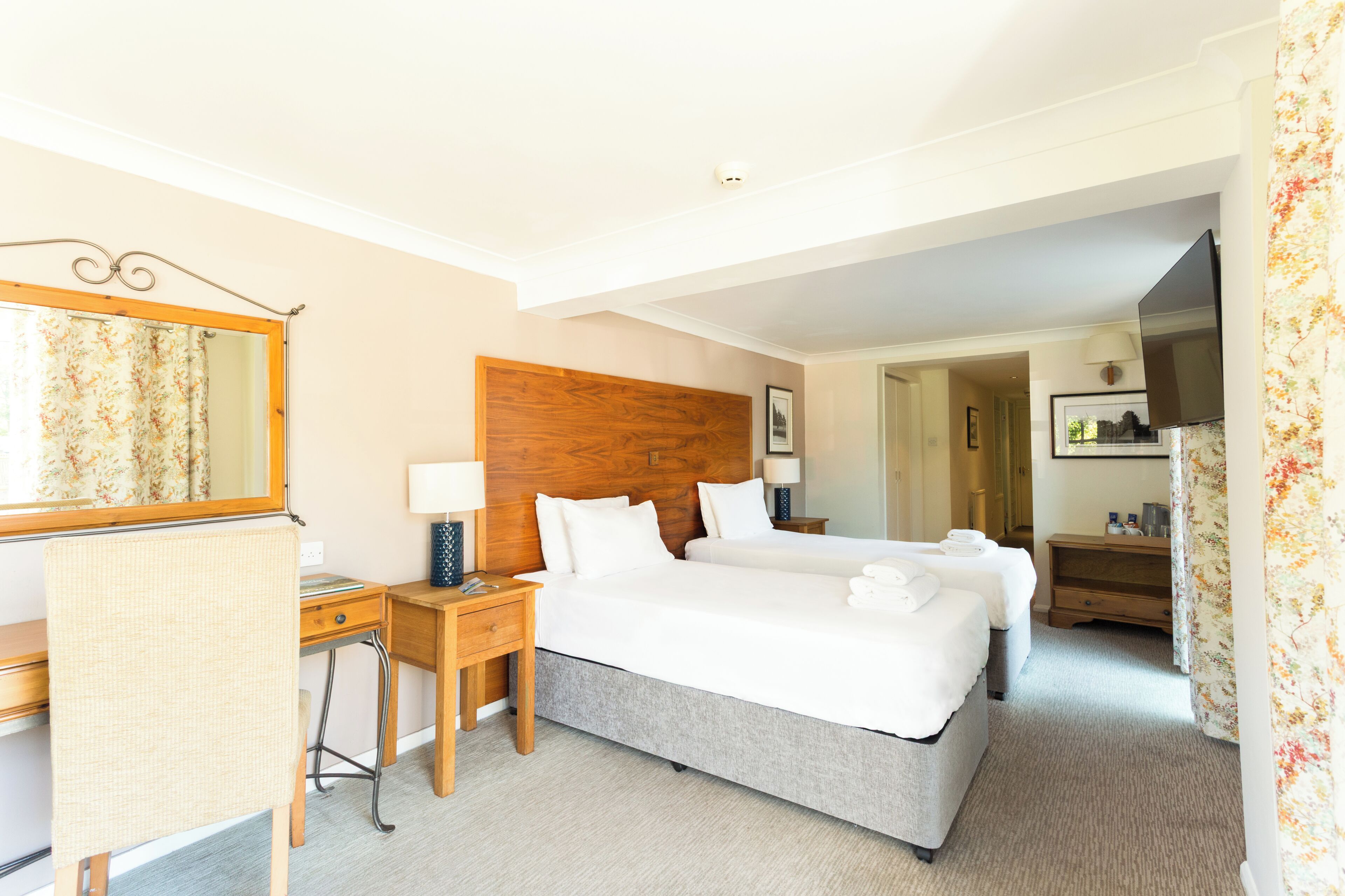 Quarto de hotel com cabeceira de madeira, duas camas, escrivaninha com cadeira, espelho e televisão.