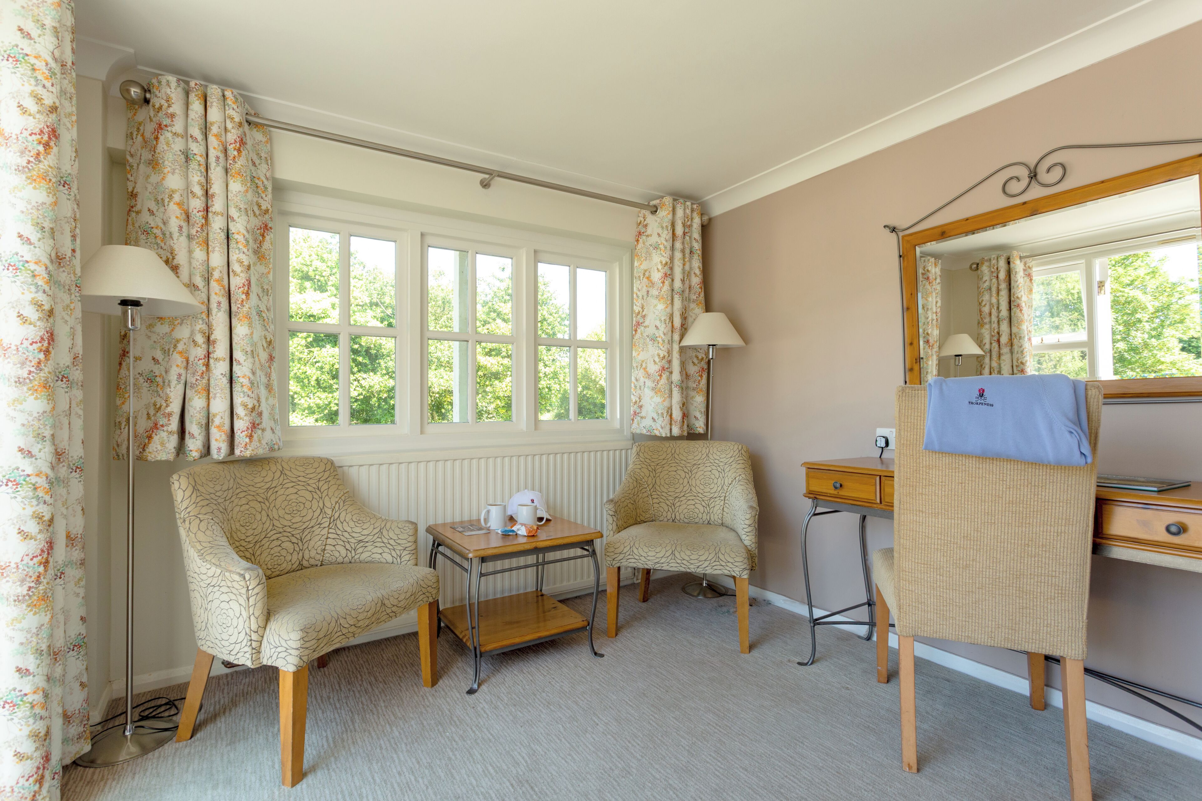 A room with two armchairs, a small table, a desk, a mirror, and a window with curtains.
