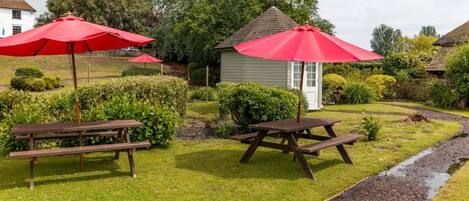 A garden with picnic tables and red umbrellas, a small shed, and a house with a thatched roof.