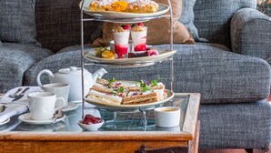 A three-tiered dessert stand with pastries and fruit on a tray, a teapot, cups, and a small bowl of jam on a table in front of a grey sofa with cushions. - Thorpeness Golf Club and Hotel (Leiston)